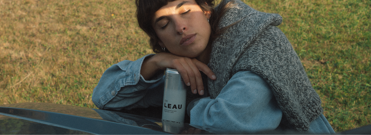 A person resting their head on a car outdoors, wearing a gray knit sweater vest over a light blue denim jacket. A silver can with the brand name 'LEAU' is resting on the car in the foreground, with dry grass in the background.