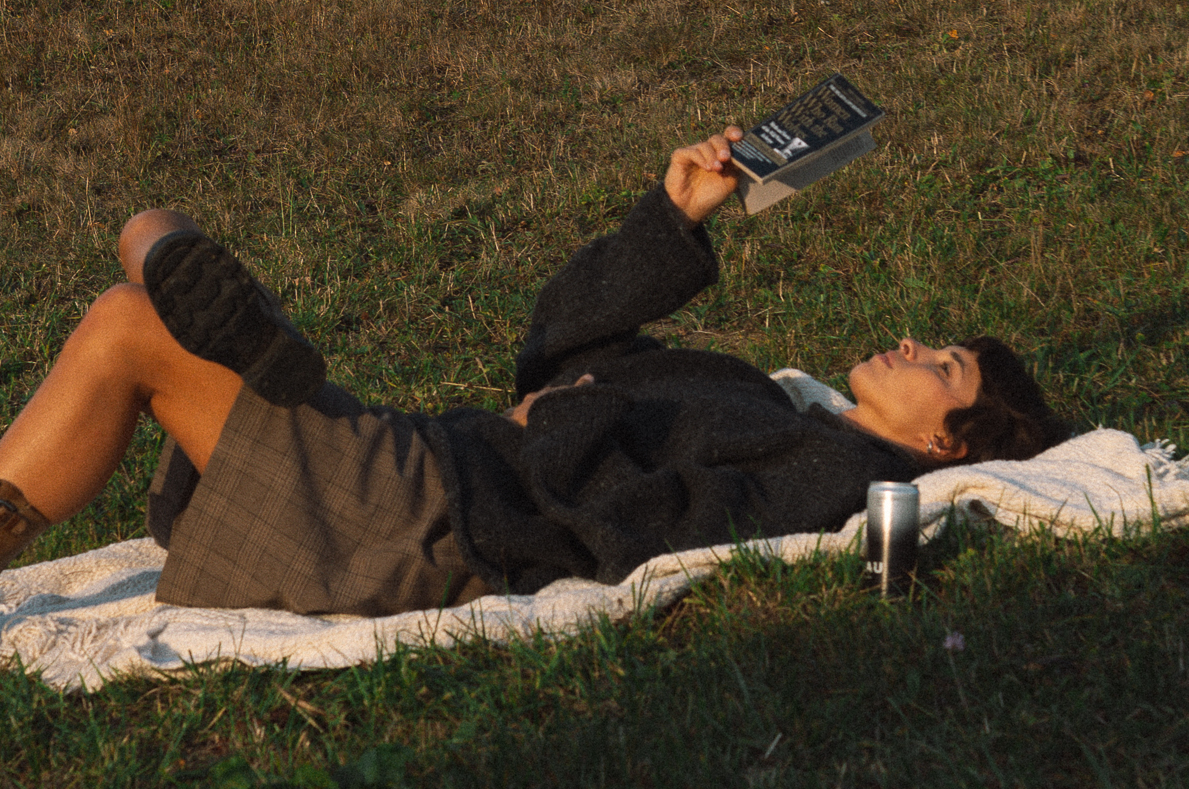 Girl laying in a field reading a book with a can of LEAU NIGHT with sunset light. 
