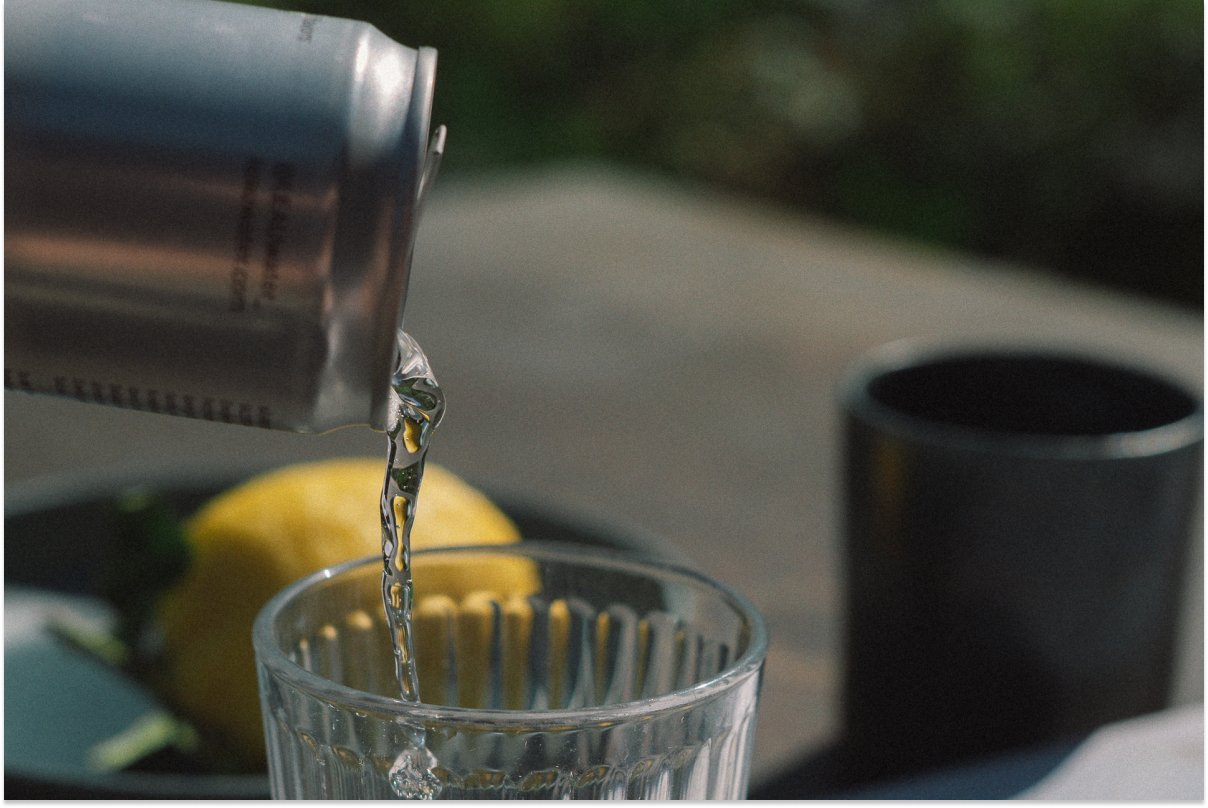 Silver can pouring water into a glass with a lemon and black cup in the background.