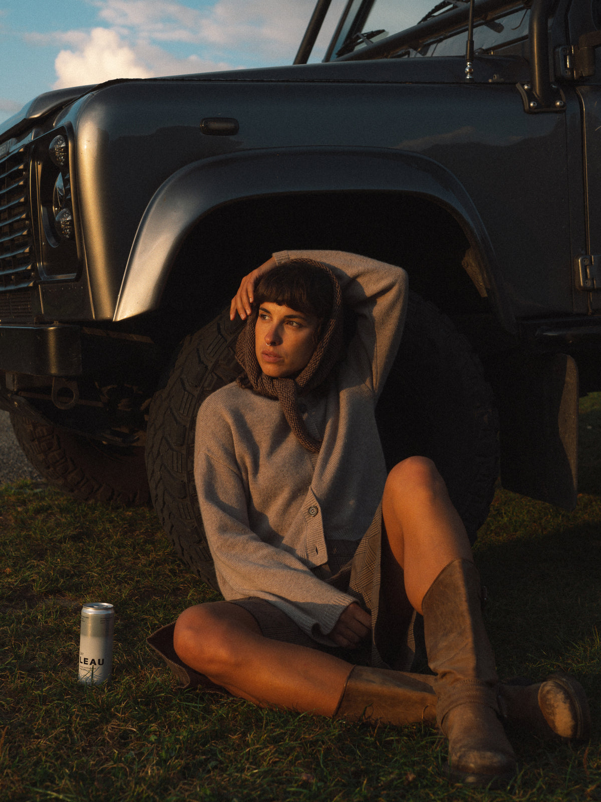 A woman sits on the grass in front of a dark off-road vehicle at sunset, wearing a light gray cardigan, brown skirt, and tall brown boots. A silver can lies on the grass nearby.