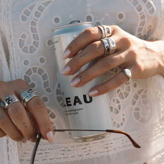 A close-up lifestyle shot of a person wearing multiple silver rings, holding a can of Leau sparkling water, with a pair of eyeglasses resting across their hands against a white lace top.