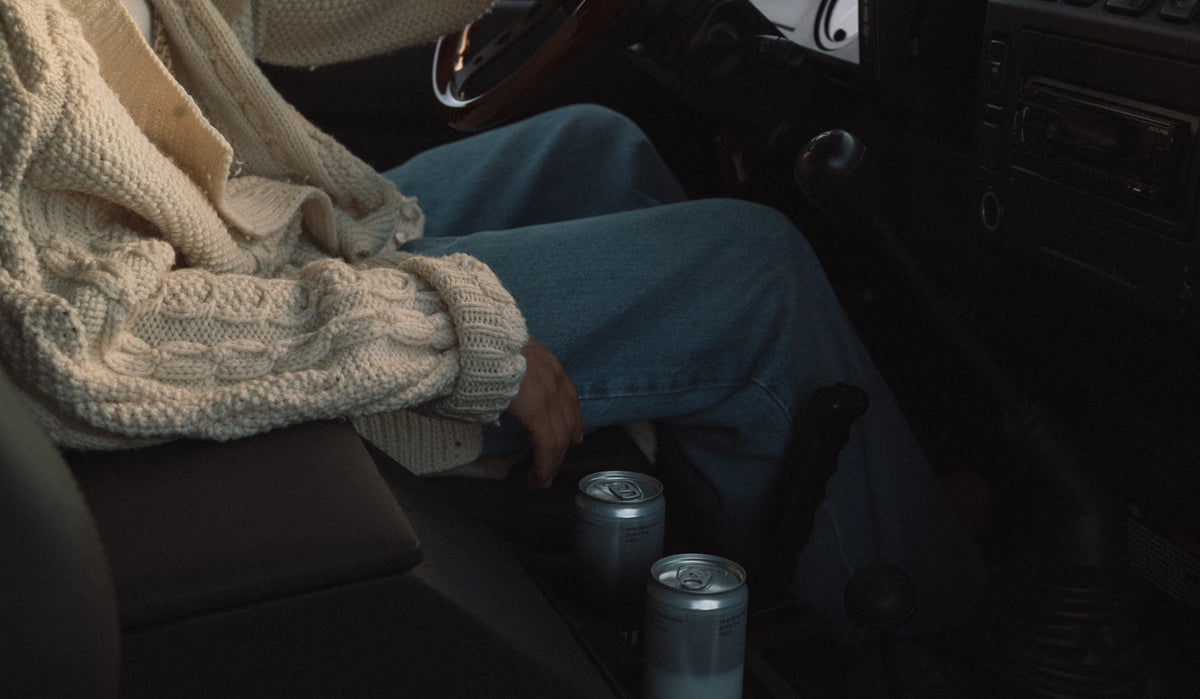 A person sitting in a car interior wearing a cream cable-knit sweater and blue jeans, with two silver aluminum cans in the cup holders.