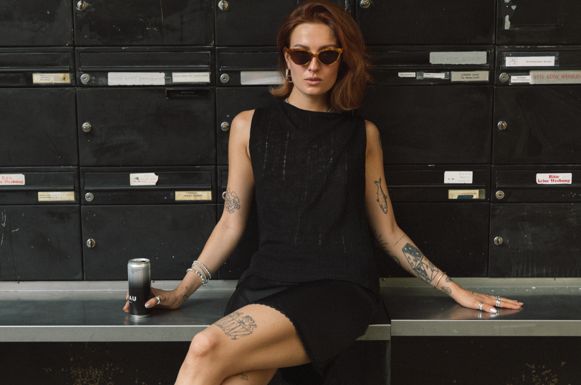 A woman with tattoos sits on a bench in front of a row of black mailboxes, wearing a sleeveless black dress and sunglasses while holding a can.