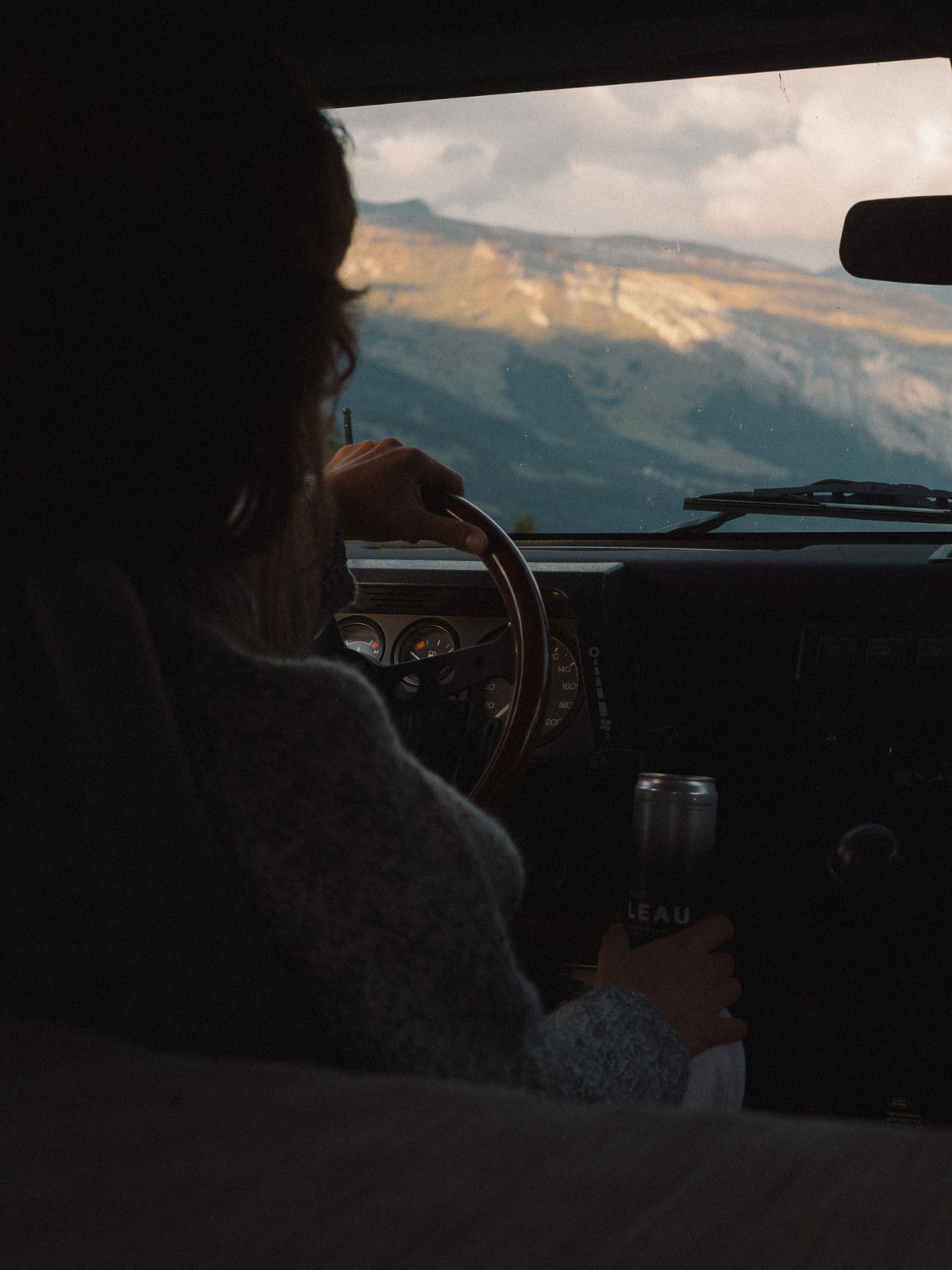 A person sitting inside a car, seen from behind, holding a can while driving past a mountain landscape visible through the windshield.