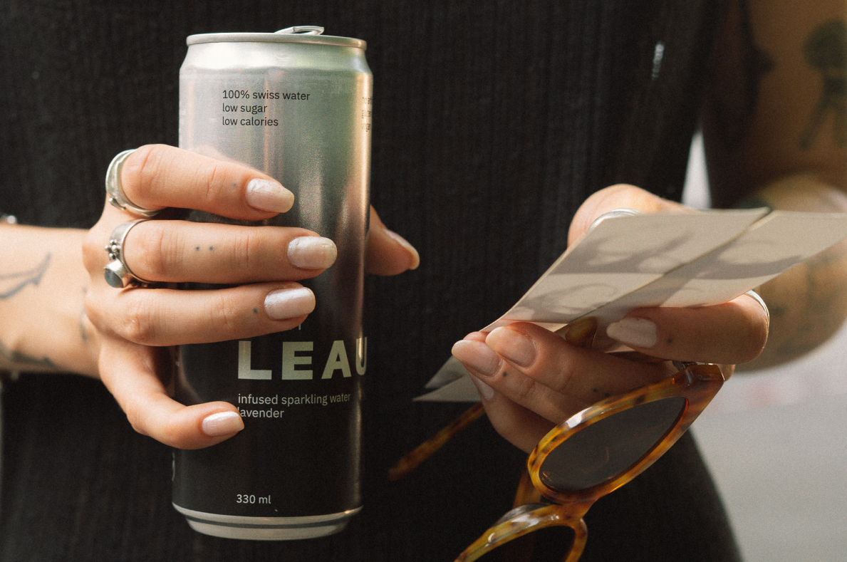 A person holding a silver can of LEAU infused sparkling water with white text, while holding a card and tortoiseshell sunglasses.