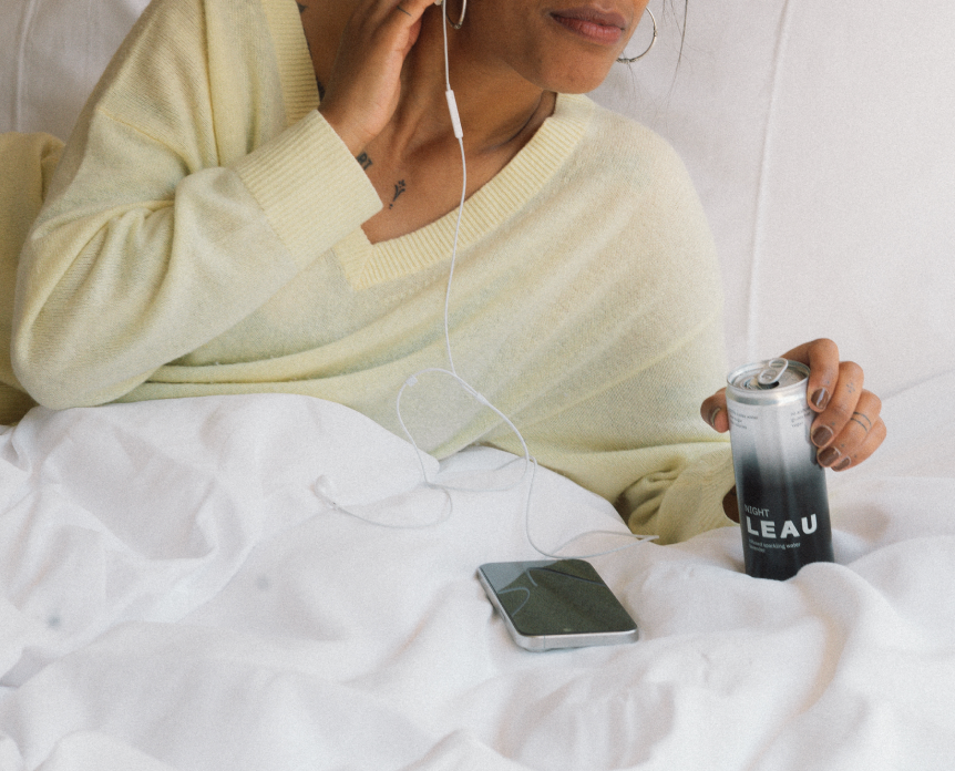 Person in bed holding a can of LEAU NIGHT and a phone, wearing headphones.