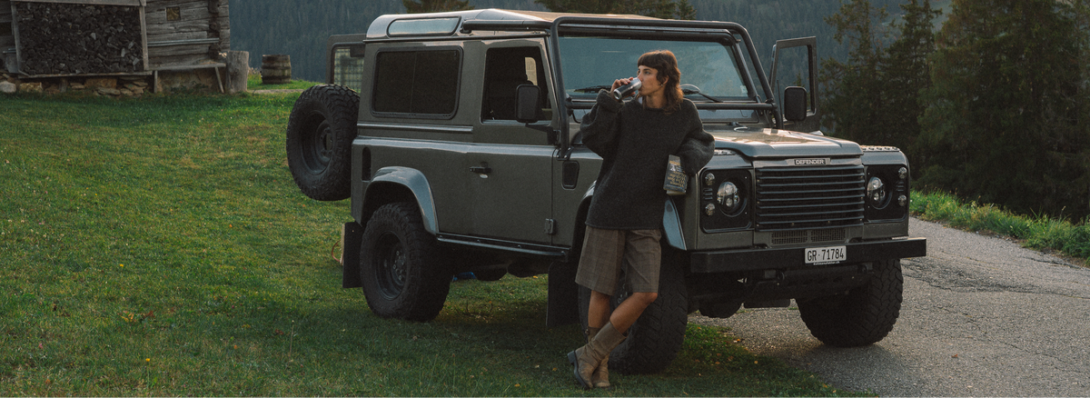 A woman leans against the open driver-side door of a dark gray off-road SUV parked on a gravel road beside a grassy field and trees.